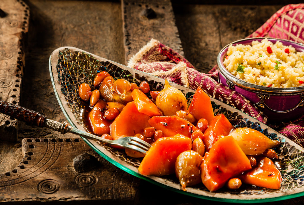 Still Life of Traditional Tajine Vegetable Dish Made with Bright Orange Vegetables in Sauce and Served in Long Narrow Modern Dish with Fork on Rustic Wooden Table with Couscous